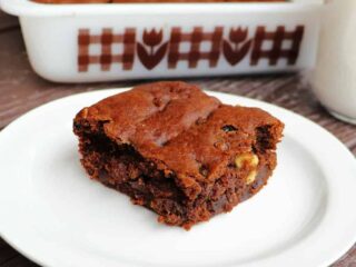 A slice of hermit bars on a white plate with a glass of milk and remaining cake pan full of bars in the background.