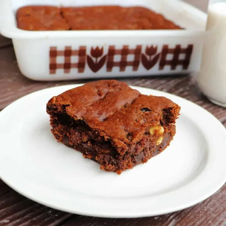 A slice of hermit bars on a white plate with a glass of milk and remaining cake pan full of bars in the background.