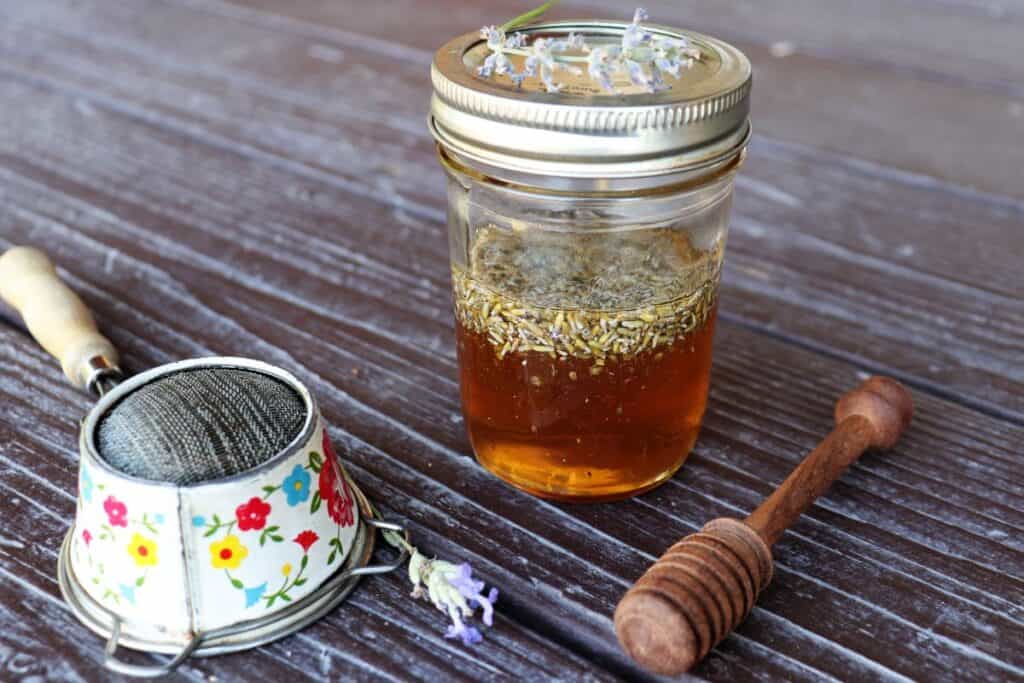 A jar of honey with lavender buds floating in it. A honey dipper, small strainer, and fresh stems of lavender surround the jar.