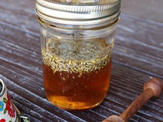 A jar of honey with lavender buds floating in it. A honey dipper and fresh stems of lavender surround the jar.