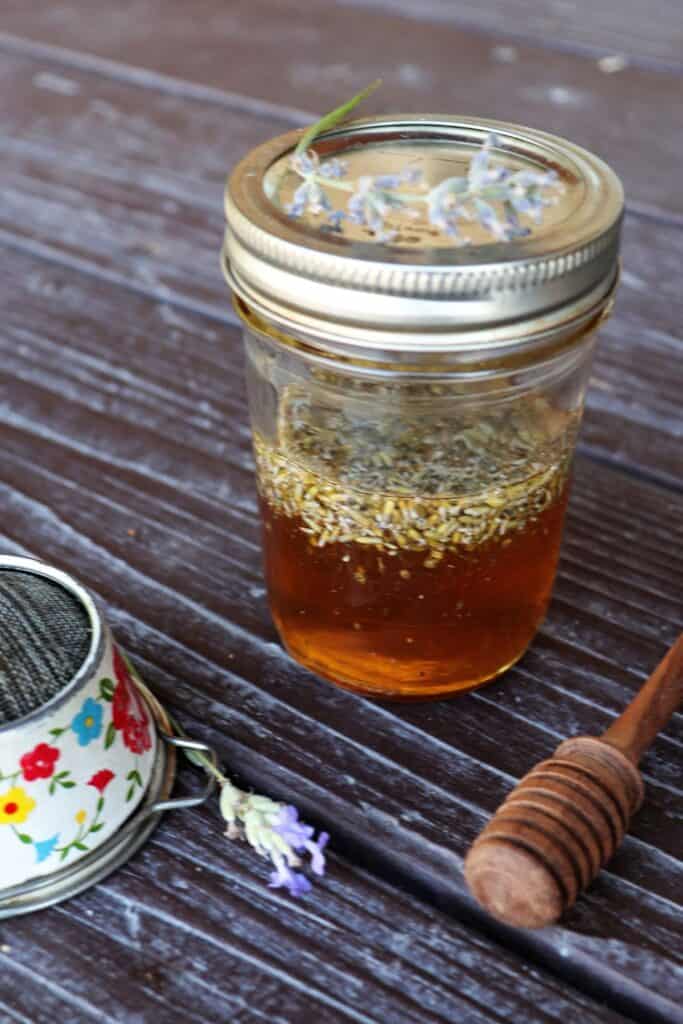 A jar of honey with lavender buds floating in it. A honey dipper, metal strainer, and fresh stems of lavender surround the jar.