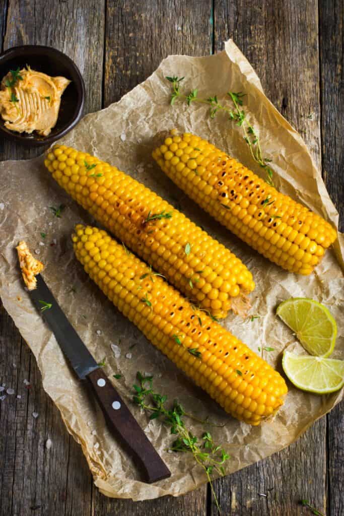 Ears of grilled corn sitting on parchment paper surrounded by fresh herbs and slices of lime.