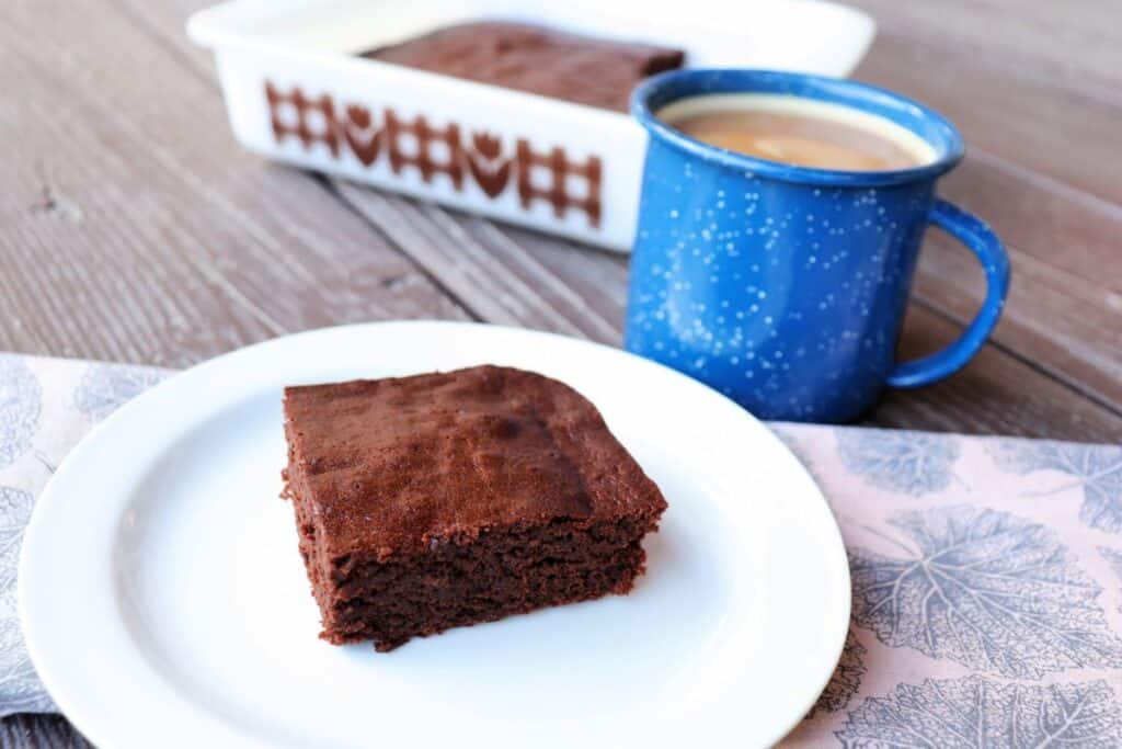 A brownie sits on plate with a blue tin cup of coffee and remaining brownies in a pan in the background. 