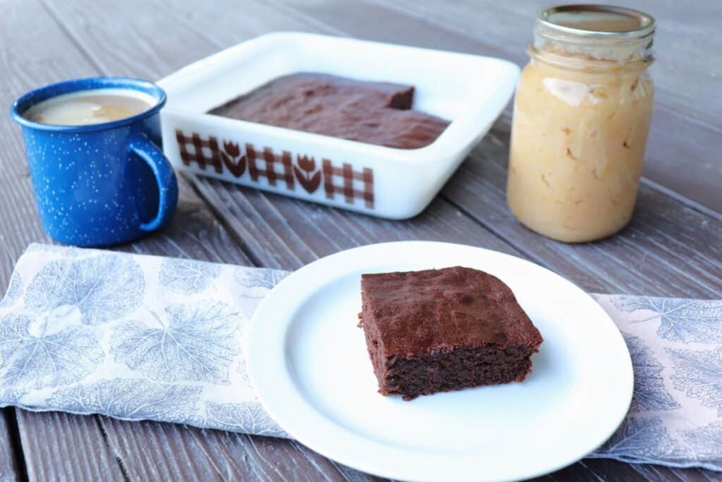 A brownie sits on a plate. In the background is a pan of the remaining brownies, a cup of coffee, and a jar of applesauce.