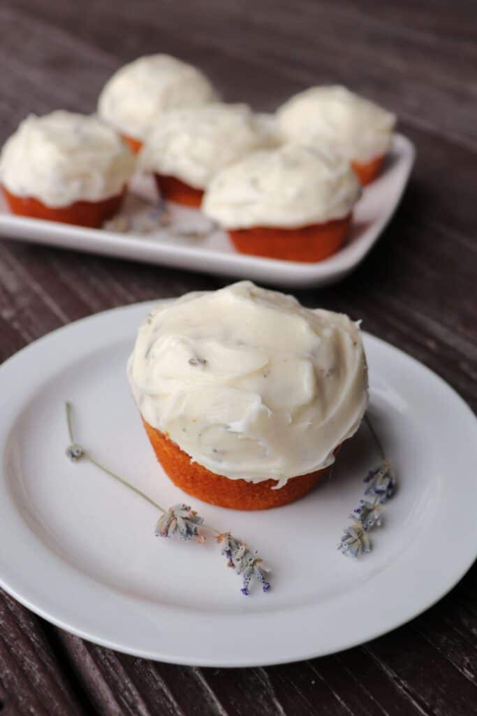 A single frosted cupcakes sits on a plate with dried stems of lavender. A platter of more cupcakes sits in the background.