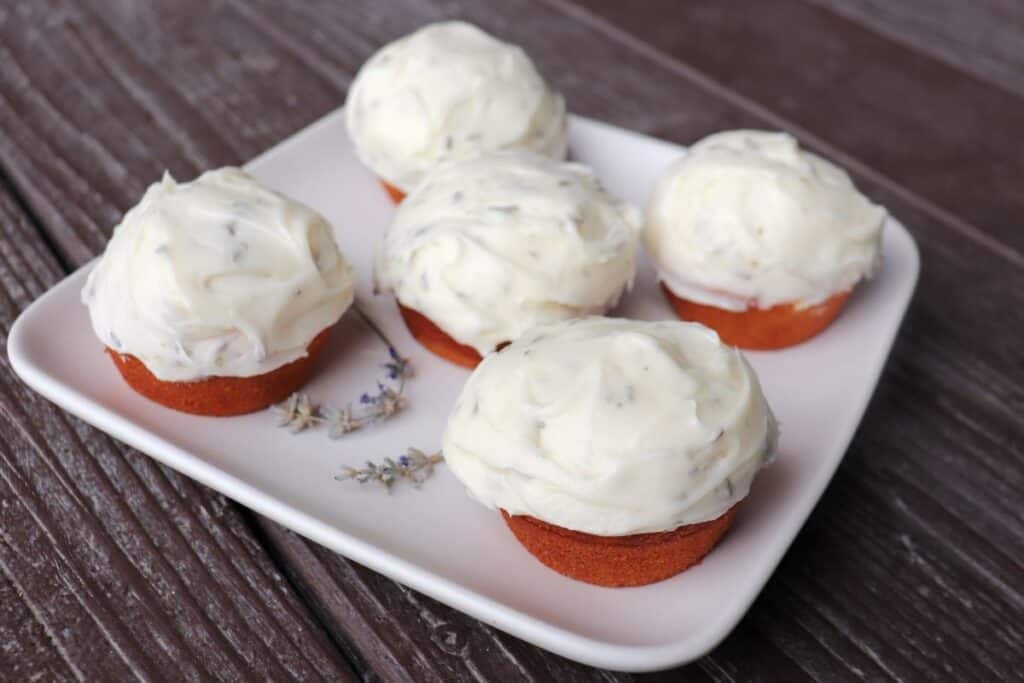 5 frosted cupcakes sit on a plate with a few stems of dried lavender. 