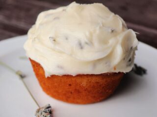A frosted cupcakes sits on a plate with a stem of dried lavender.