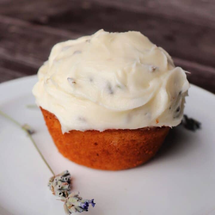 A frosted cupcakes sits on a plate with a stem of dried lavender.
