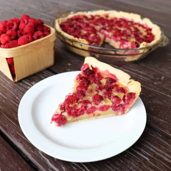 A slice of raspberry custard pie sits on a plate with the remaining whole pie in the background and a box of fresh raspberries.