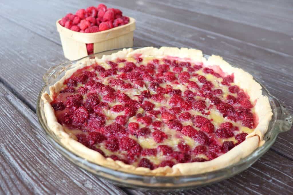 A whole raspberry custard pie in a glass pie plate sits on a table with a box of fresh raspberries behind it. 
