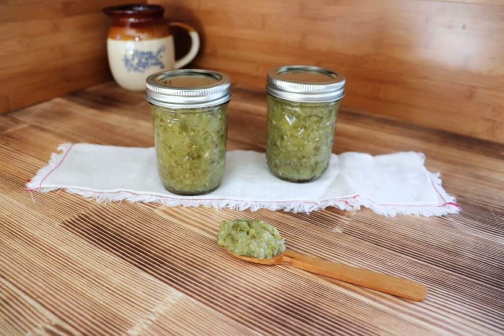 Two jars of relish sit on top of a white cloth with a wooden spoon with relish sitting on top in front of them. 