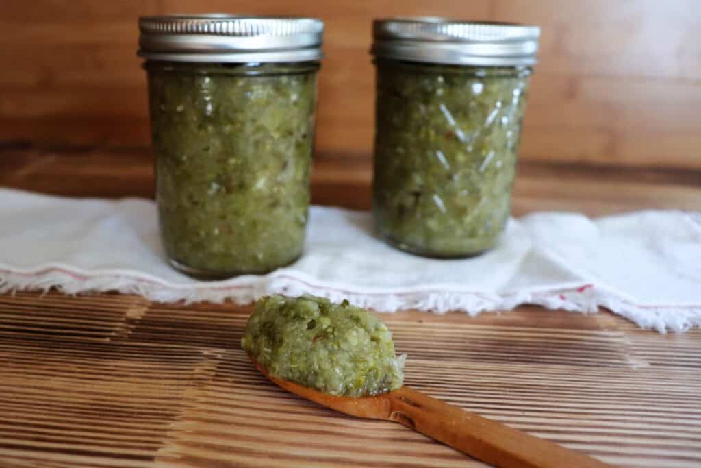 Two jars of relish sit on top of a white cloth with a wooden spoon with relish sitting on top in front of them. 