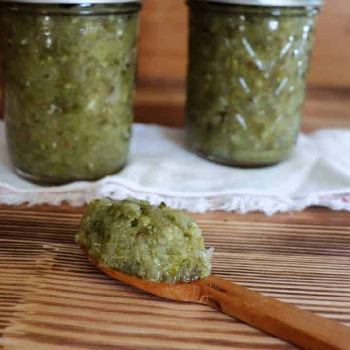 A small wooden spoon with dill pickle relish on top sits in front of 2 jars of relish sitting on a white cloth.