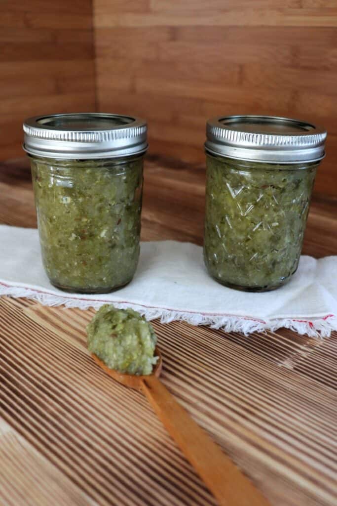 Two jars of relish sit on top of a white cloth with a wooden spoon with relish sitting on top in front of them. 