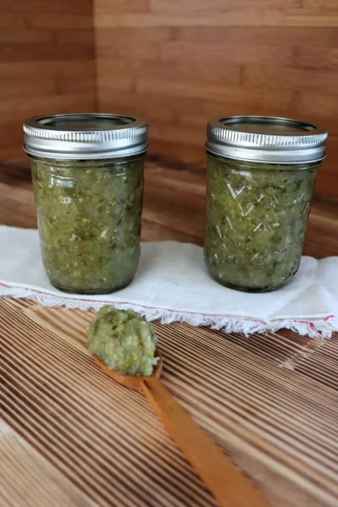 Two jars of relish sit on top of a white cloth with a wooden spoon with relish sitting on top in front of them. 