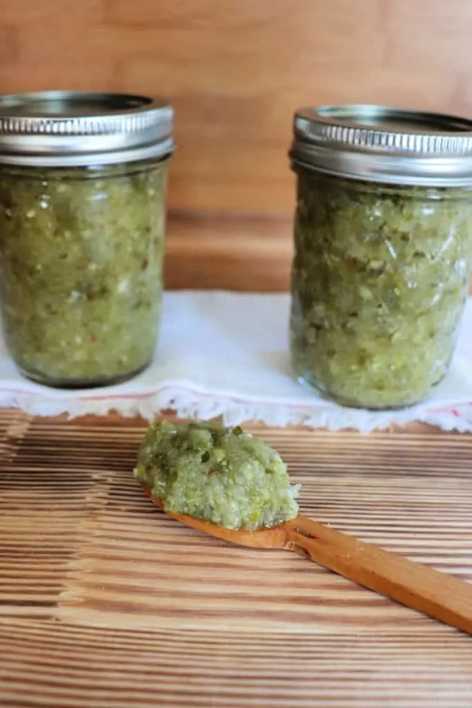 Two jars of relish sit on top of a white cloth with a wooden spoon with relish sitting on top in front of them. 