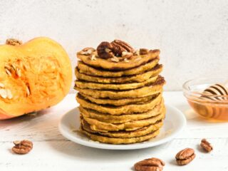 A stack of pancakes sit on a plate with a cut pumpkin and bowl of honey in the background.