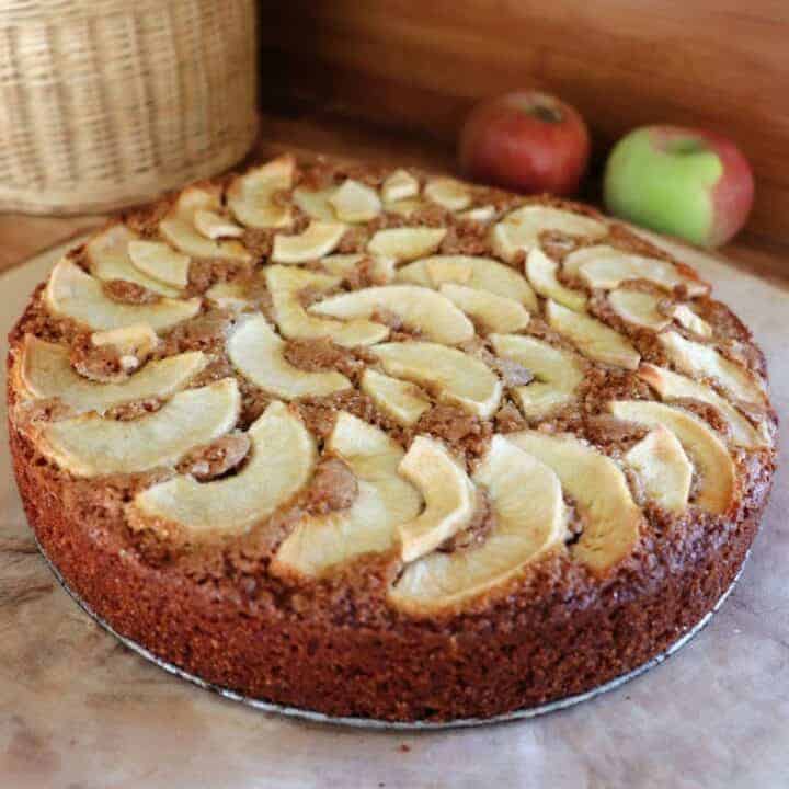 An apple cake sits on a board with fresh apples in the background.