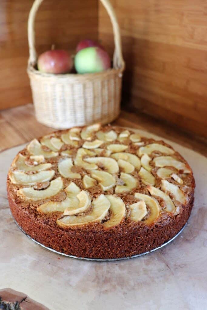 A whole apple cakes sits on a board with a basket of fresh apples in the background. 