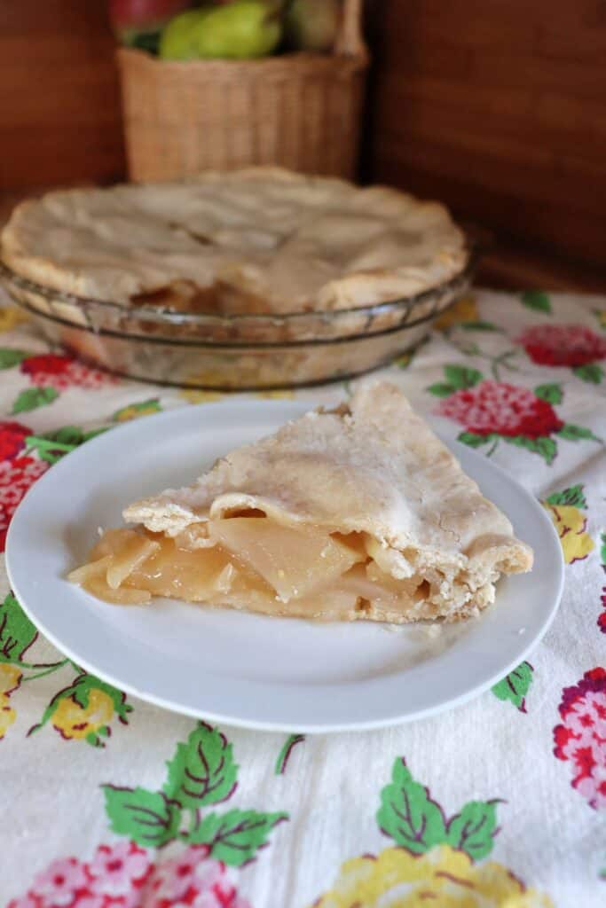 A slice of pie sits on a plate with remaining pie behind it. In the background is a basket full of fresh apples and pears. 