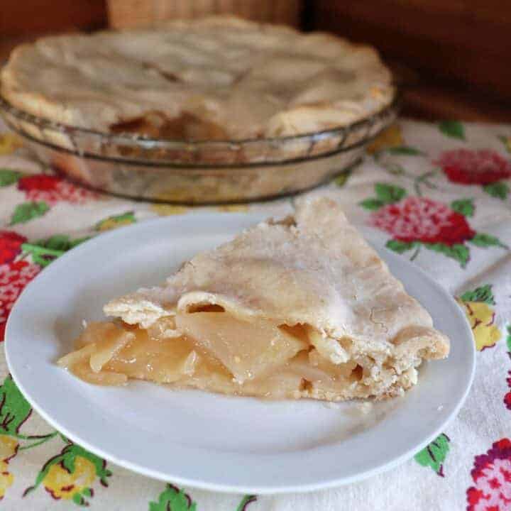 A slice of apple pear pie sits on a plate. Remaining dish of pie in the background.
