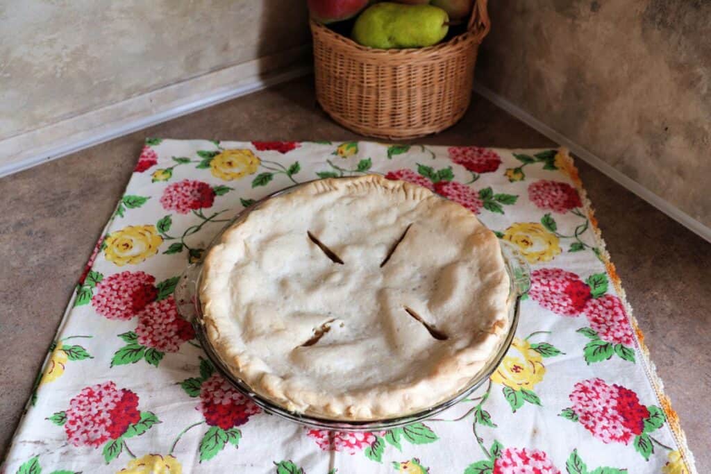 A whole pie sits on a top of a floral cloth with a basket of fresh pears in the background.