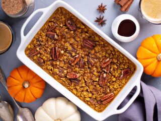 A pan full of baked oatmeal with chopped nuts as seen from above is surrounded by pumpkins and whole spices.