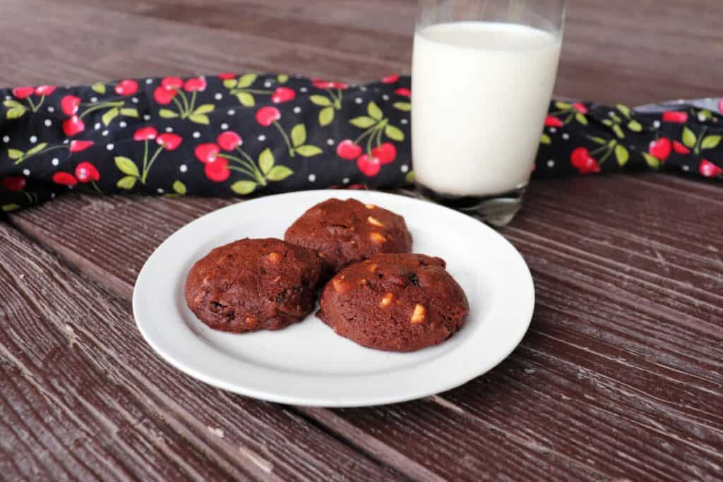 3 cookies on a plate with a cherry covered cloth and glass of milk in the background.