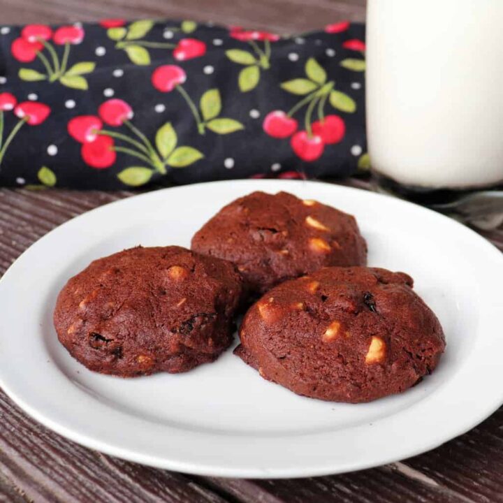 3 cookies on a plate with a glass of milk and a cherry decorated cloth in the background.