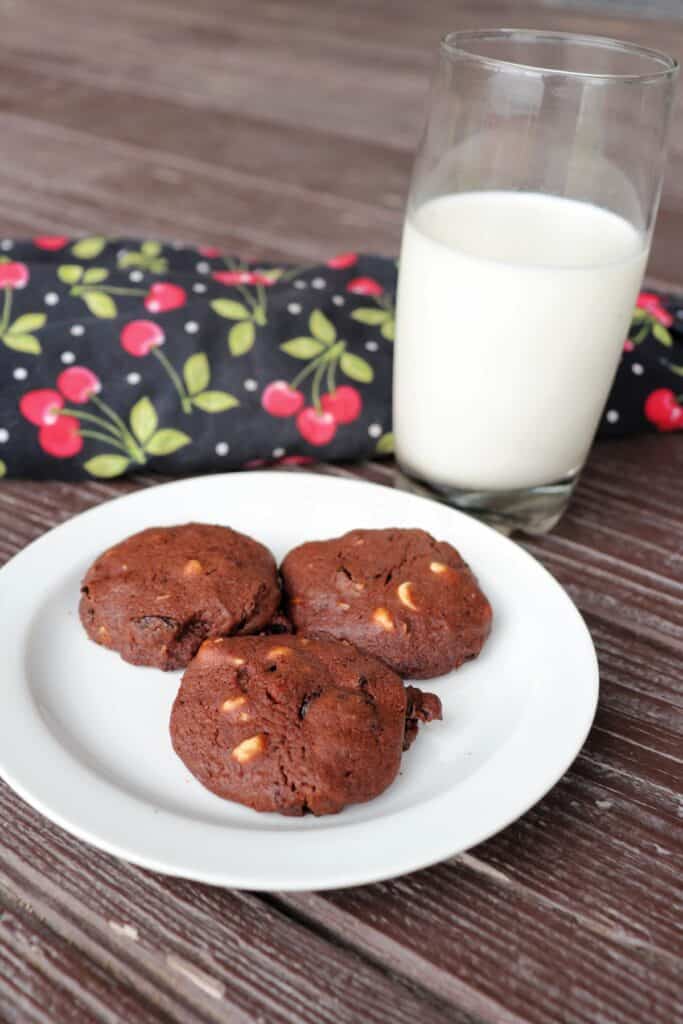 3 cookies on a plate with a cherry covered cloth and glass of milk in the background.