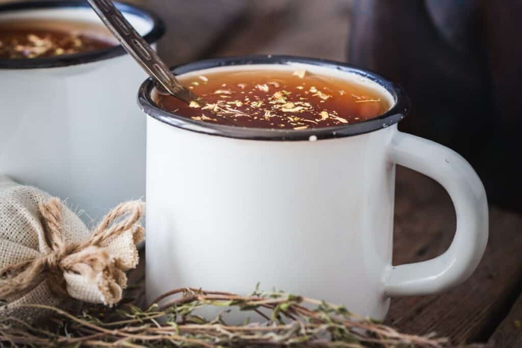 A white enamel cup full of tea with herbs floating on top, a spoon sticks out of the cup.