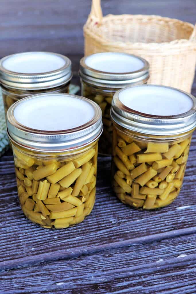 4 canning jars full of green beans sit on a table with a basket in the background. 