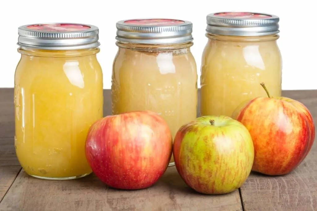 3 jars of applesauce lined up in front of fresh apples. 