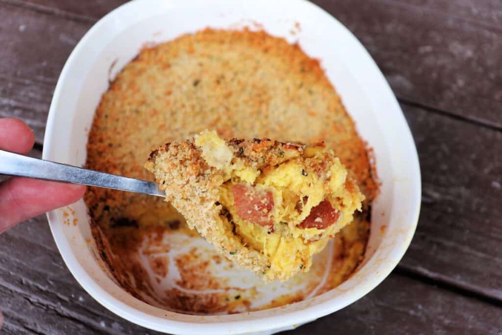 A spoonful of acorn squash casserole showing bits of bacon being held above the remaining casserole dish.