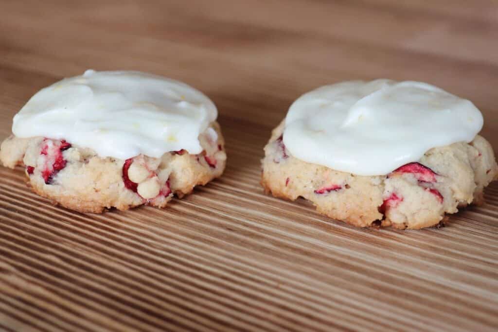2 frosted cranbery cookies sit next to each other on a wooden board. 