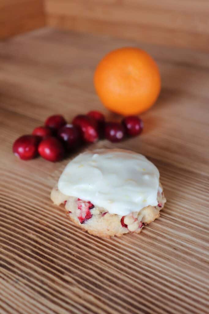 A frosted cranberry cookie sits on a board with fresh cranberries and an orange behind it. 