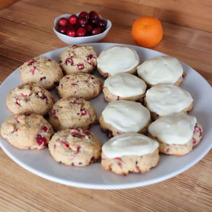 Cranberry orange cookies sit on a plate, some have frosting, some do not. Behind the plate is a bowl of fresh cranberries and and an orange.
