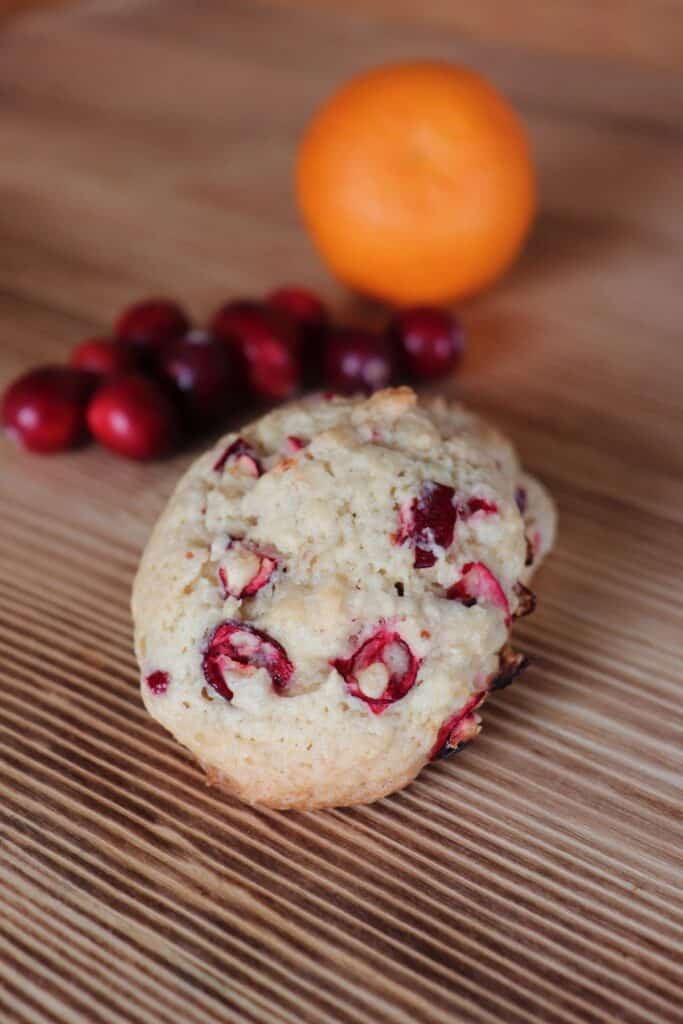 A cookie sits on a board showing up bits of chopped cranberries cooked inside. Fresh cranberries and an orange sit behind it. 