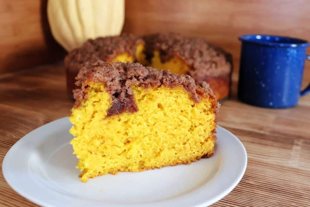 A slice of orange colored pumpkin coffee cake with a streusel topping sits on a white plate with a blue coffee cup and the remaining cake in the background. 