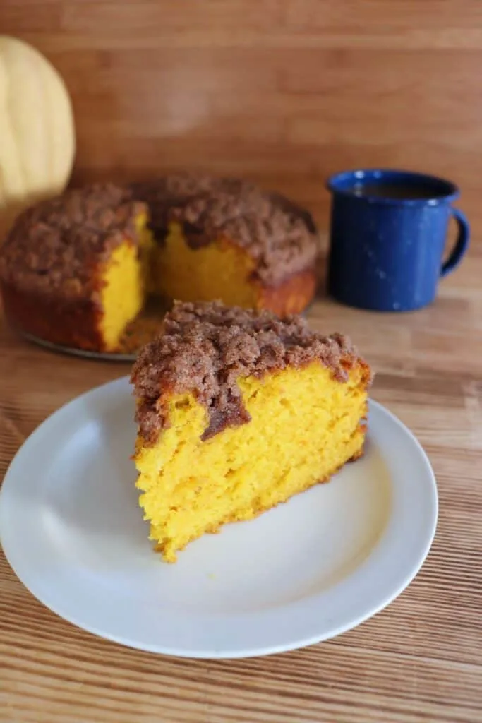 A slice of orange colored pumpkin coffee cake with a streusel topping sits on a white plate with a blue coffee cup and the remaining cake in the background. 