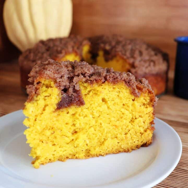 A slice of pumpkin coffee cake with streusel topping sits on a white plate with the rest of the cake and a winter squash in the background.