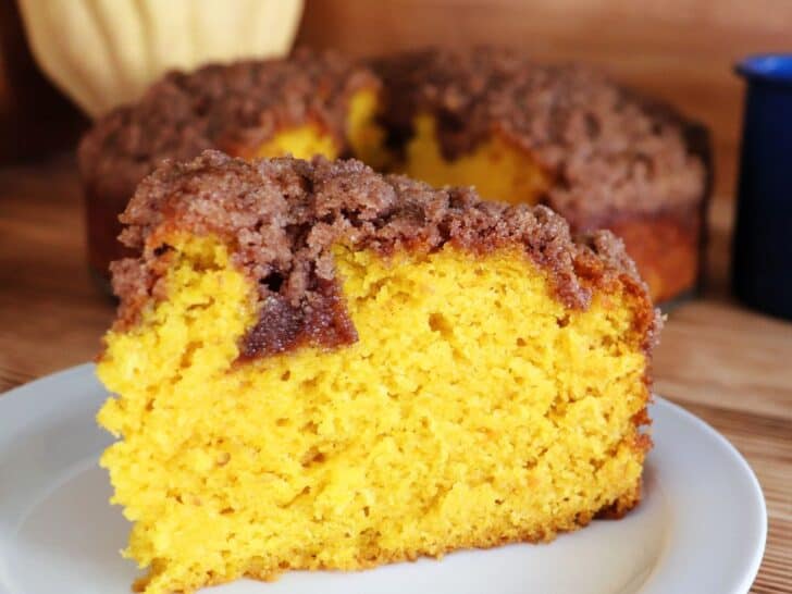 A slice of pumpkin coffee cake with streusel topping sits on a white plate with the rest of the cake and a winter squash in the background.