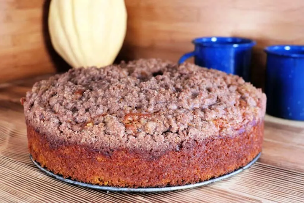 A whole pumpkin coffee cake with streusel topping sits on a table. In the background are two blue coffee cups and an acorn squash. 