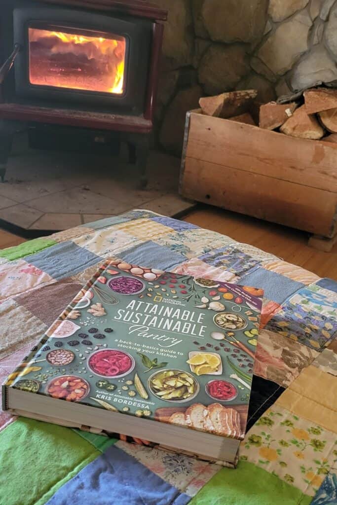 A hardcovered book titled, 'Attainable Sustainable Pantry' sits on an ottoman covered in a colorful patchwork quilt. In the background is a fire is burning inside a red woodstove.