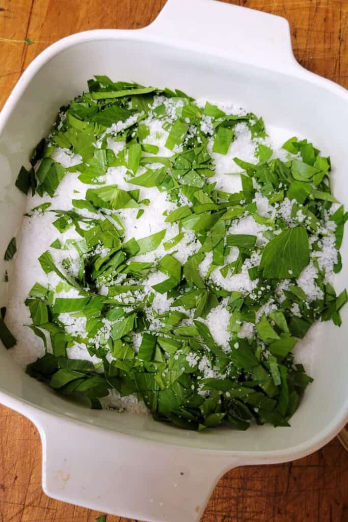 Fresh green lovage leaves are chopped an mixed with salt in a small, white baking dish. 