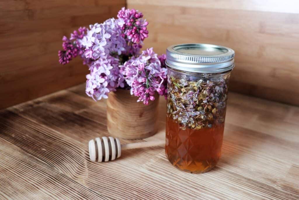 A glass canning jar full of honey and lilac blossoms sits on a table. Behind it is a wooden vase full of fresh lilac blooms. A wooden honey dipper sits between the jar and vase.