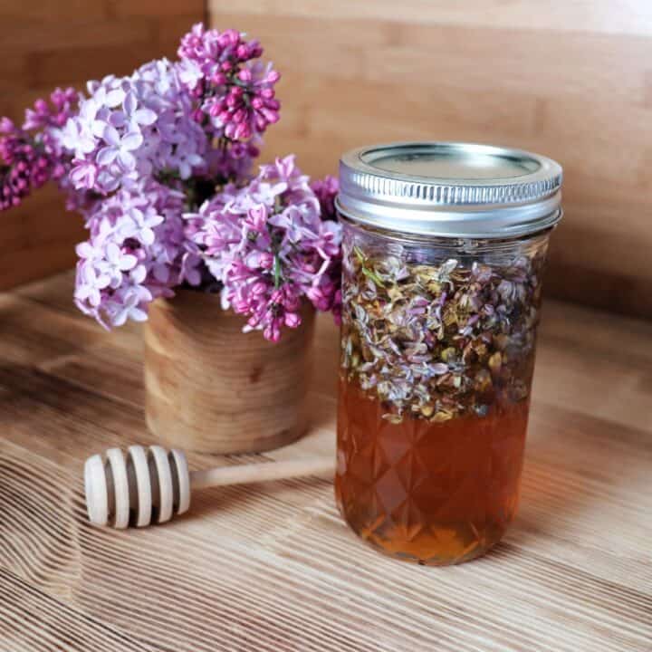 A glass canning jar full of honey and lilac blossoms sits on a table. Behind it is a wooden vase full of fresh lilac blooms. A wooden honey dipper sits between the jar and vase.