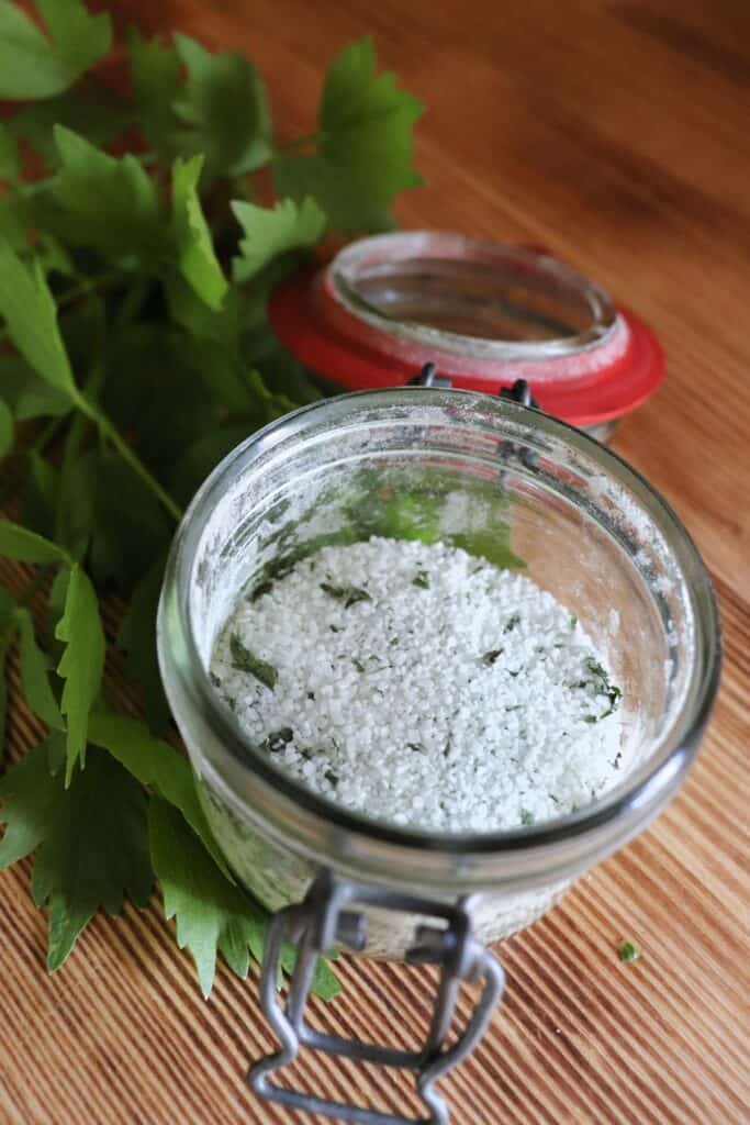 An open glass jar sits on table, inside the jar is salt and dried herbs all mixed together. next to the jar is a stalk of fresh lovage leaves. 