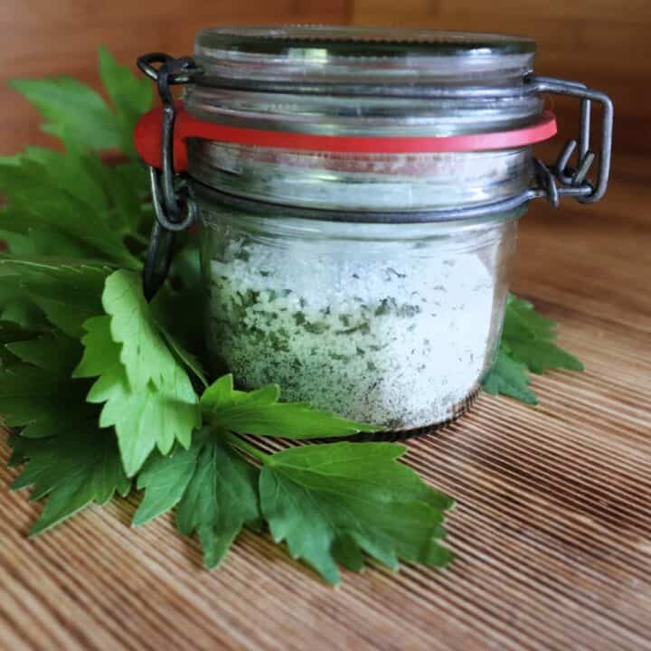 A small jar full of salt and dried herbs sits on a table next to some fresh lovage leaves.