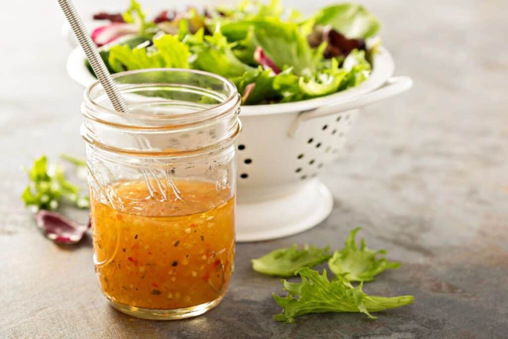 A glass jar full of salad dressing and a whisk sticking out of it sits on a table in front of a colander full of lettuce.